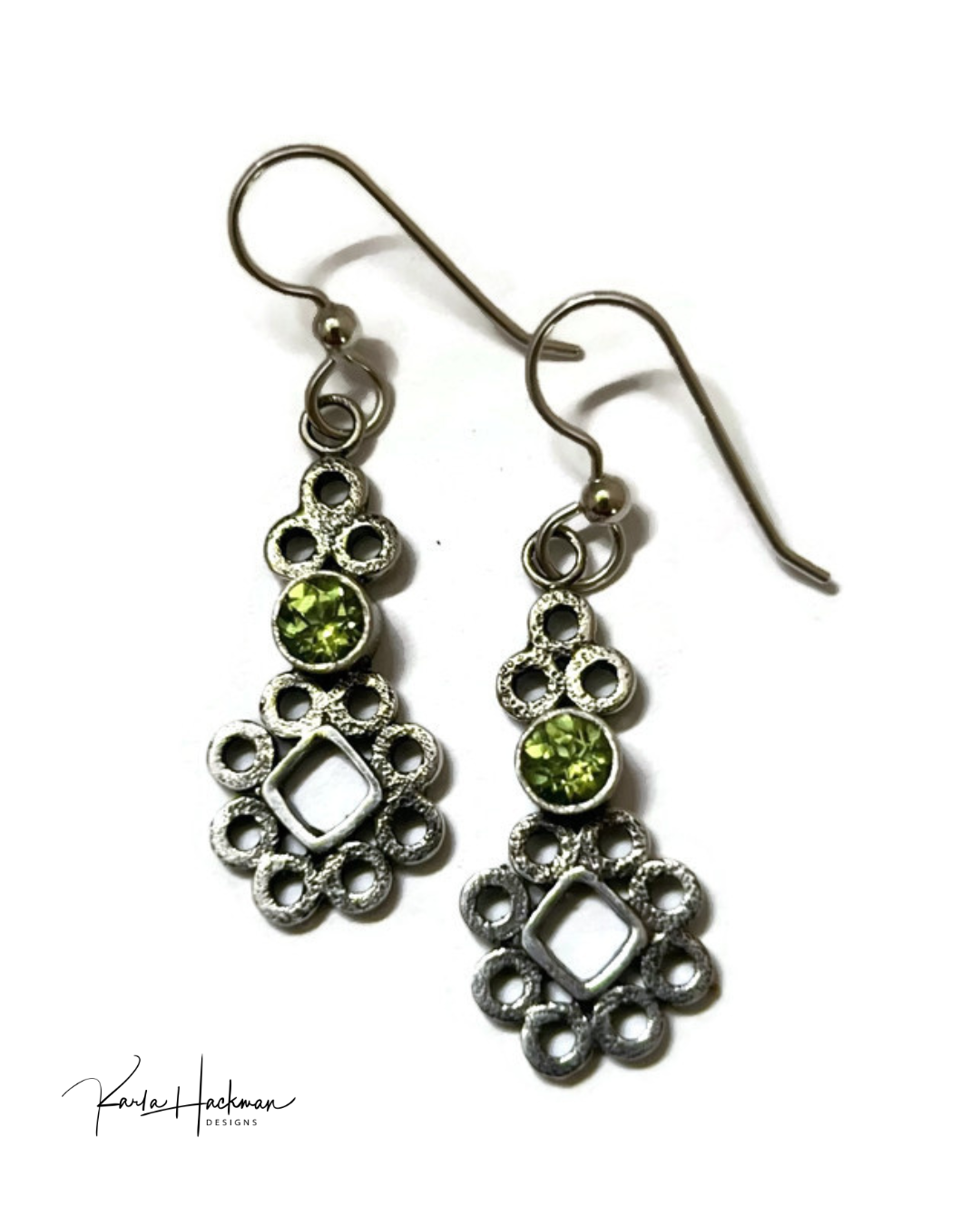 A pair of silver-toned dangle earrings with green peridot accents, photographed on a plain white background. A signature watermark for &